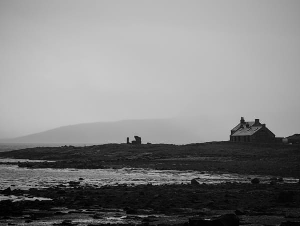 Black and white image of a rocky beach with two cottages on the right and the ruins of a building central.